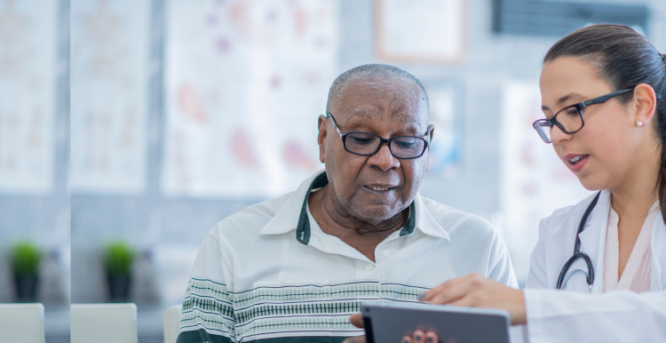 A doctor in a white coat and stethoscope shows a patient something on a tablet