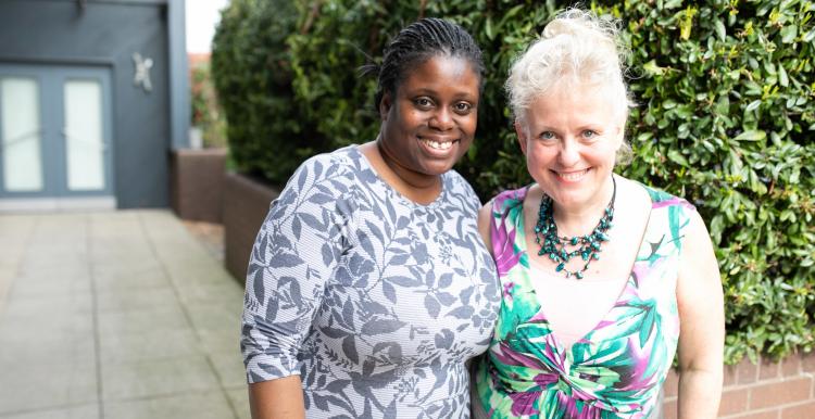 Two women standing in front of a hedge with their arms around each other and smiling at the camera