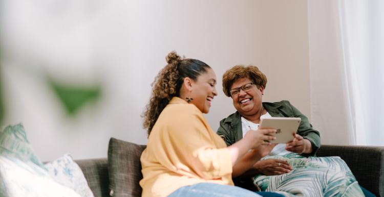 Two women, one older and one younger, sit next to each other in a bright room on a sofa and laugh while looking at a tablet. Both have dark skin and curly hair.