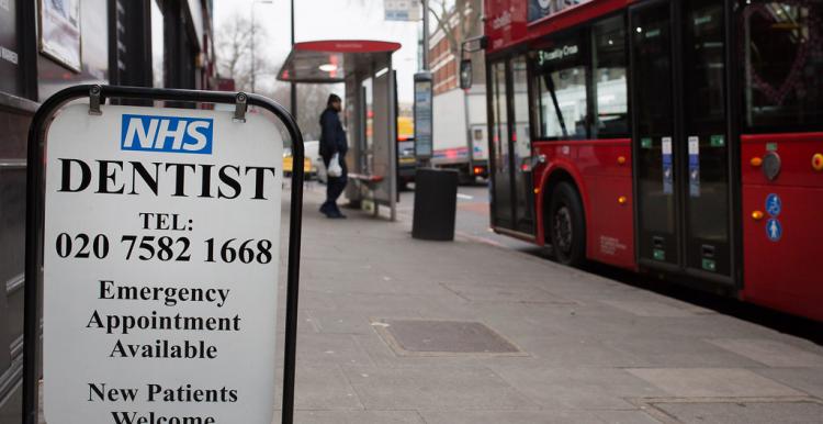 A photograph of a sign on the street advertising an NHS dentist with a London telephone number and emergency appointments. The street behind the sign is blurred, but a covered bus stop and red double decker bus are visible.