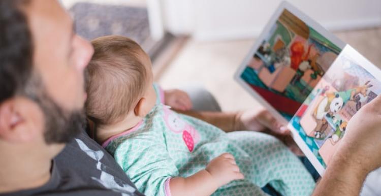 A photograph taken from above of a father and baby looking at a picture book together. The baby is wearing a blue and pink patterned onesie. .