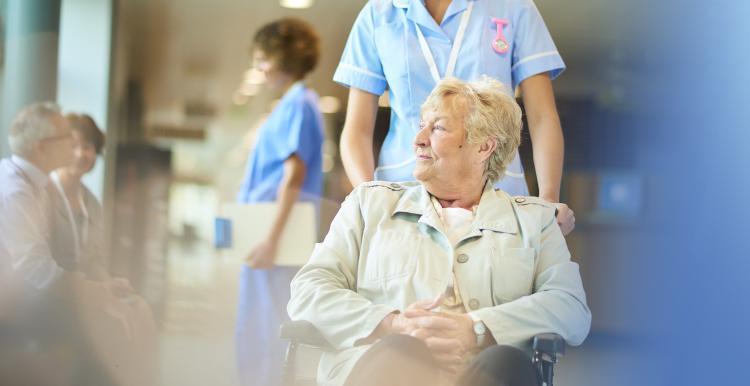 A patient in a wheelchair is pushing down a hospital corridor by a nurse.