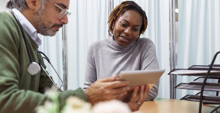 A black female patient reviews medical test results on a tablet with her male doctor of Middle Eastern descent. They are seated next to each other at a desk. 