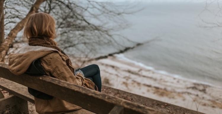 A woman viewed from behind sitting on a bench overlooking a beach on a cold, grey day.