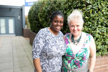 Two women standing in front of a hedge with their arms around each other and smiling at the camera