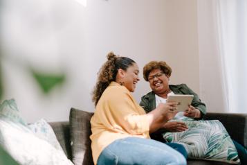 Two women, one older and one younger, sit next to each other in a bright room on a sofa and laugh while looking at a tablet. Both have dark skin and curly hair.