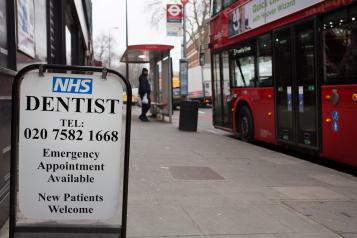 A photograph of a sign on the street advertising an NHS dentist with a London telephone number and emergency appointments. The street behind the sign is blurred, but a covered bus stop and red double decker bus are visible.