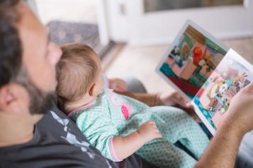 A photograph taken from above of a father and baby looking at a picture book together. The baby is wearing a blue and pink patterned onesie. .