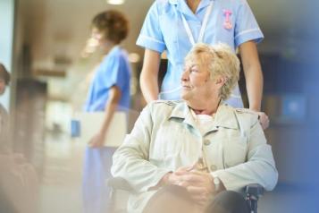 A patient in a wheelchair is pushing down a hospital corridor by a nurse.