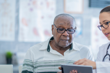 A doctor in a white coat and stethoscope shows a patient something on a tablet