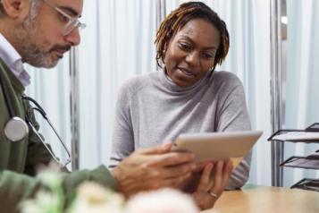 A black female patient reviews medical test results on a tablet with her male doctor of Middle Eastern descent. They are seated next to each other at a desk.