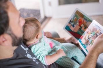 A baby sits on their father's lap while he reads from a picture book. The baby is wearing a light blue patterned onesie. The photo is taken from an angle looking over the father's shoulder from above.  