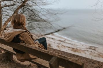 A woman viewed from behind sitting on a bench overlooking a beach on a cold, grey day.