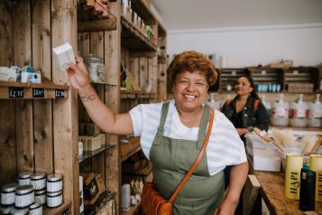 A smiling woman with brown skin and short hair reaches for a box on a shelf in a shop.  