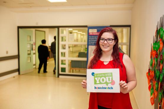 Woman stood in a hospital entrance holding a sign that says "It starts with you" and the Healthwatch logo