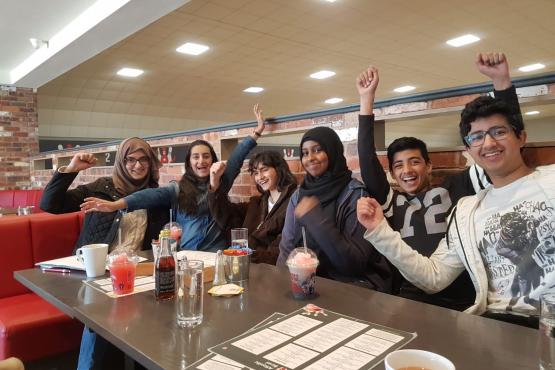 A group of smiling young volunteers sit at a cafe table cheering and raising their arms. The group is made of up multiple genders, ethnicities, and ages. 