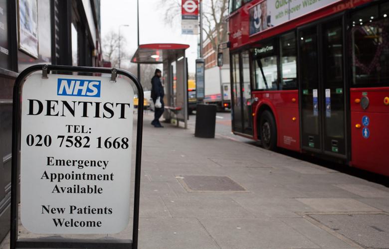 A photograph of a sign on the street advertising an NHS dentist with a London telephone number and emergency appointments. The street behind the sign is blurred, but a covered bus stop and red double decker bus are visible.