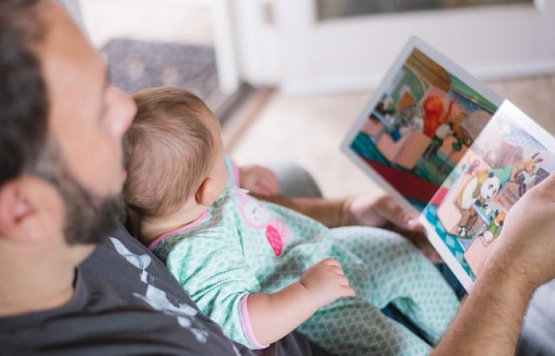 A photograph taken from above of a father and baby looking at a picture book together. The baby is wearing a blue and pink patterned onesie. .