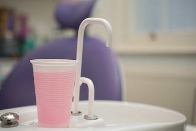 A photograph of a white plastic cup full of pink mouthwash on a small sink in a dental practice. The cup is in focus, and the office in the background is blurred.