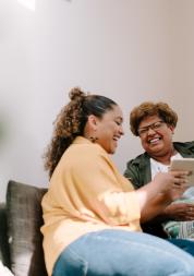 Two women, one older and one younger, sit next to each other in a bright room on a sofa and laugh while looking at a tablet. Both have dark skin and curly hair.
